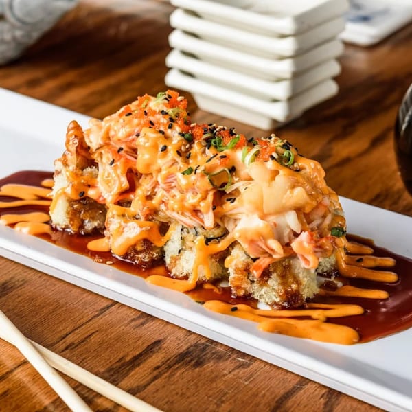 Crispy sushi roll topped with spicy crab, orange roe, sesame seeds, and sauce on a white plate.